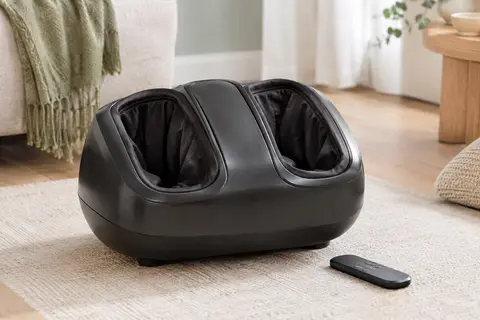 Masseur de pieds électrique noir avec télécommande, posé dans un intérieur clair aux tons sauge et bois.
