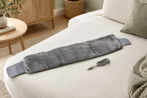 Coussin chauffant lombaire gris en peluche, déplié sur une assise claire avec ses attaches velcro visibles.